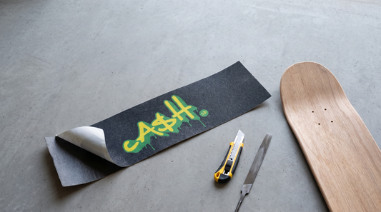 Lija de skate CASH negra despegada de su papel, junto a una tabla de maple, un cuchillo cartonero y una lima